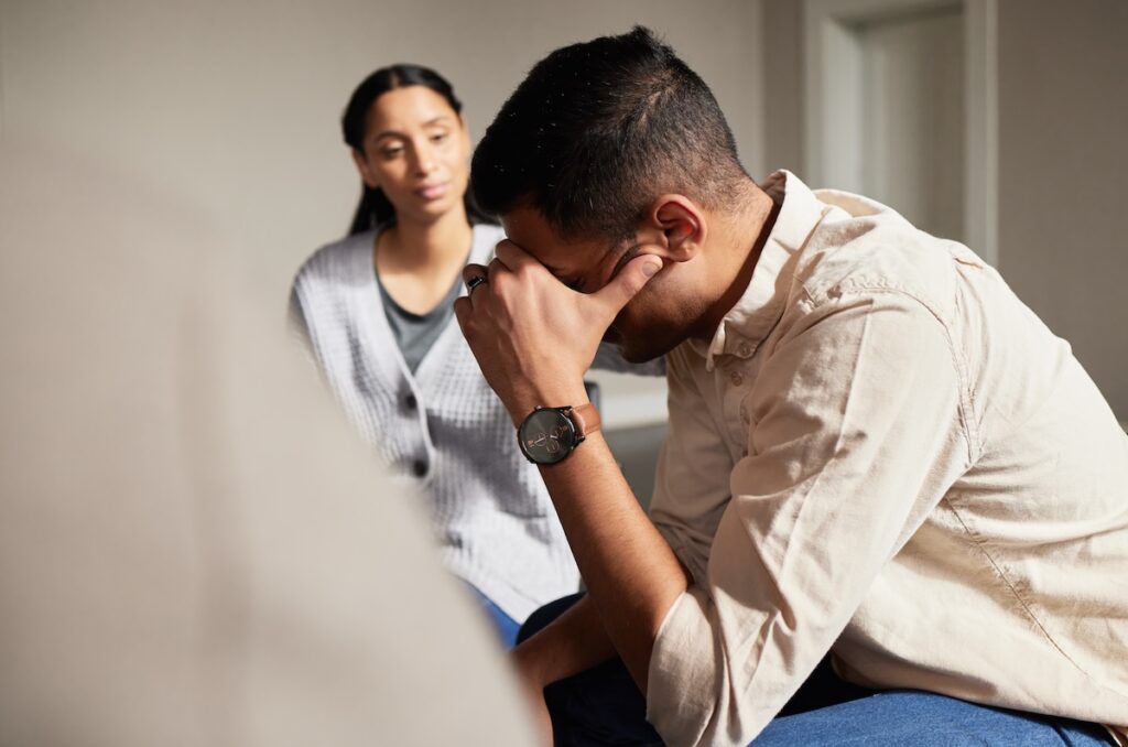 A supportive scene showing a man sitting with his hand on his forehead while another person listens attentively, offering emotional support.