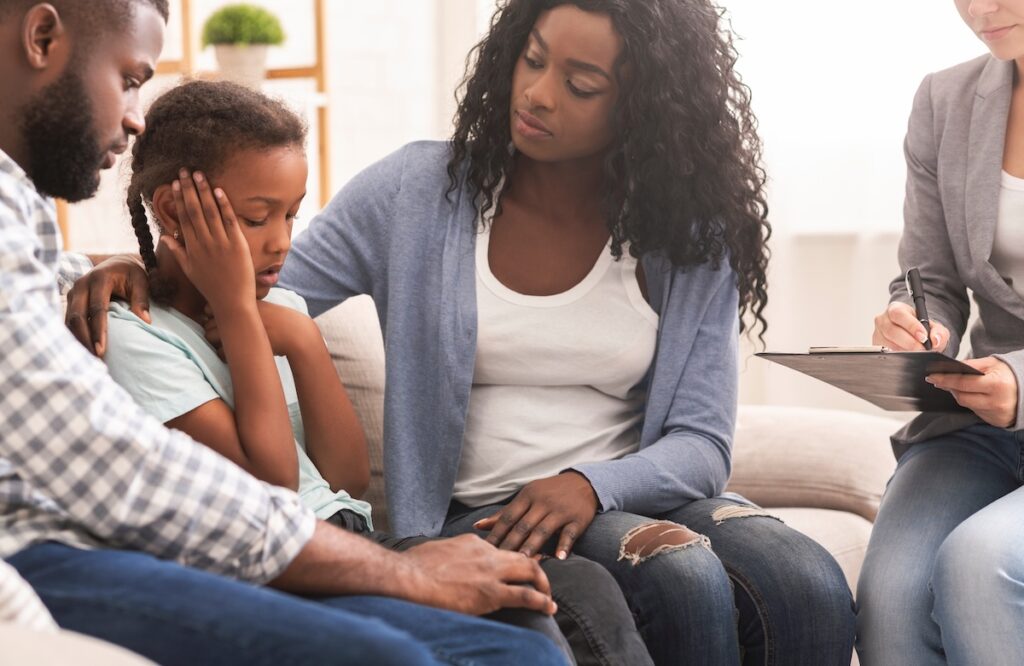 Parents comfort their distressed daughter as a therapist takes notes nearby.