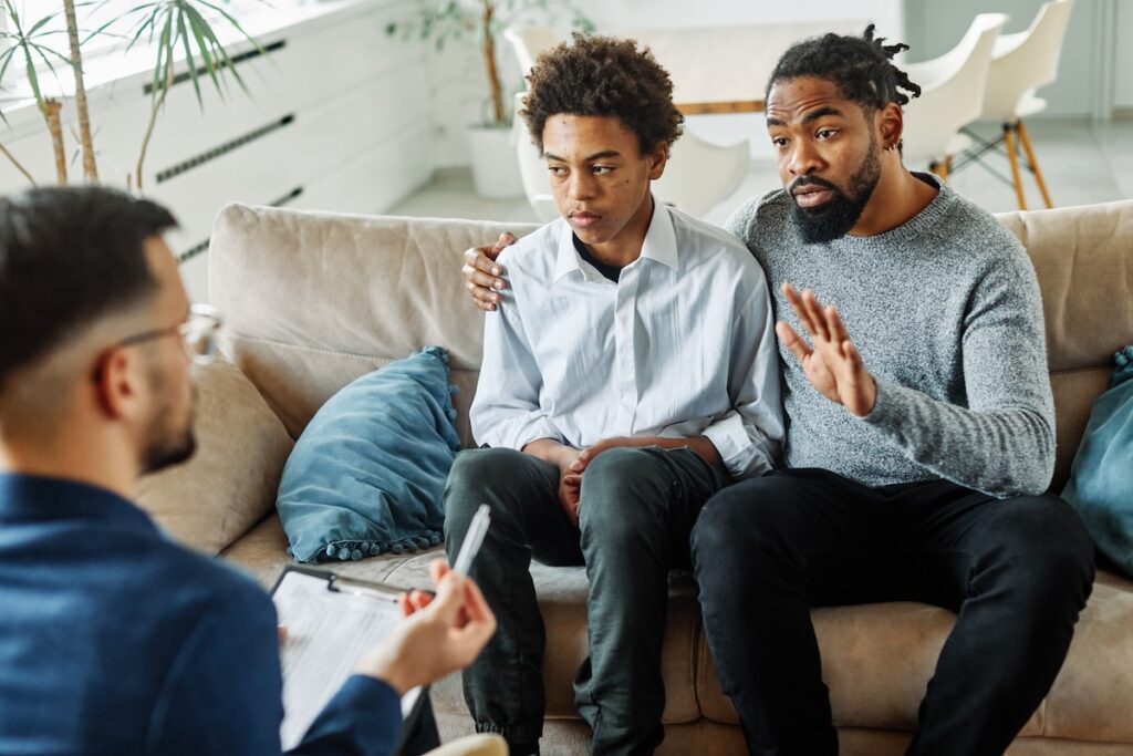 A father and son sit together on a couch during a therapy session, talking with a counselor.