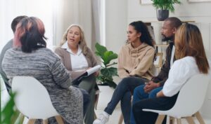 A diverse group of people sitting in a circle having a discussion during a therapy or support group session in a bright, comfortable room.
