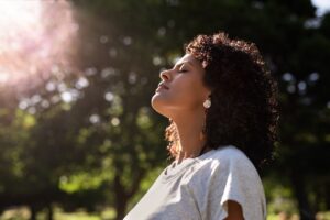 Woman standing outdoors with her eyes closed and face tilted toward the sunlight, appearing calm and peaceful, surrounded by blurred green trees in the background.