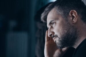 Close-up of a man with gray hair and a beard resting his head on his hand, gazing thoughtfully out of a window with a serious, pensive expression in dim lighting.