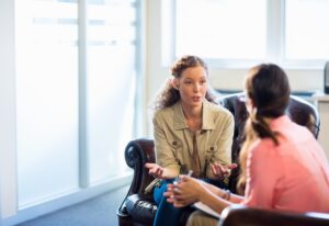 A woman sitting and talking with a therapist in a comfortable office setting, expressing herself with open hand gestures while the therapist listens attentively.