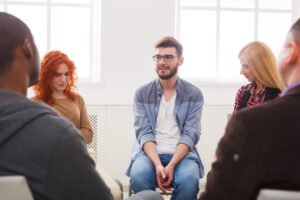 Group therapy session with several people sitting in a circle, where a man in glasses and casual clothing is speaking while others listen attentively.