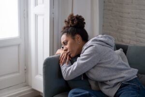 Young woman in a gray hoodie lying on a couch and staring out the window, looking sad and emotionally overwhelmed.