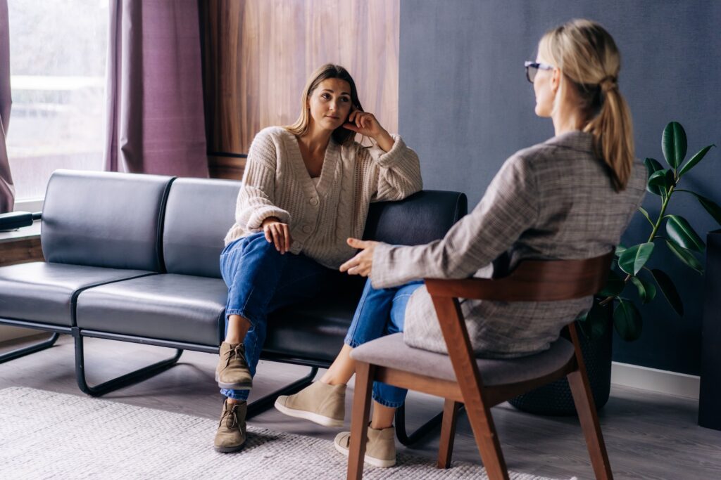 A woman sitting on a couch talking with a therapist in an office, appearing thoughtful and engaged in conversation during a counseling session.