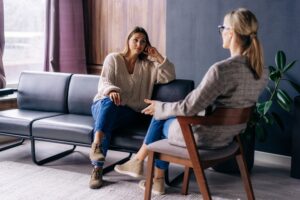 A woman sitting on a couch talking with a therapist in an office, appearing thoughtful and engaged in conversation during a counseling session.