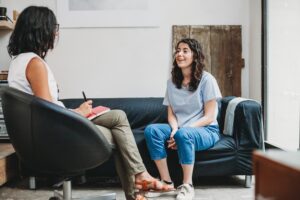 A young woman sitting on a couch smiling and talking with a therapist who is taking notes during a counseling session in a cozy, well-lit office.