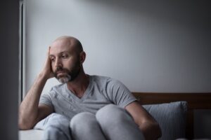 Bald man with a beard sitting on a bed near a window, resting his head on his hand and looking down with a sad, contemplative expression.