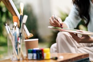 A person sits outdoors holding a paint palette and using a brush to mix colors, with jars of paint and brushes visible in the foreground.