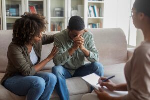 A man sits on a couch leaning forward with his hands clasped near his face, appearing distressed, while a woman beside him gently touches his arm in support. Another person sits across from them holding a clipboard, suggesting a counseling or therapy session.