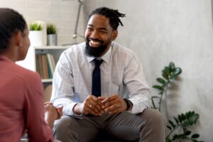 A man in a dress shirt and tie sits and smiles warmly while talking with another person in the foreground, creating a friendly and supportive atmosphere, possibly in a counseling or mentoring setting.