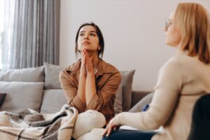 A young woman sits on a couch with her hands pressed together under her chin, looking upward with a distressed or contemplative expression, while another woman, possibly a therapist, sits nearby listening attentively.