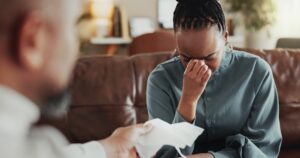 A woman sits on a couch holding her face in distress, appearing to cry, while another person in the foreground reaches out to offer her a tissue.