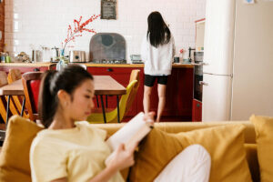 One woman reading on couch while her sober living roommate washes the dishes